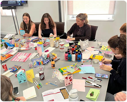 A group of people sitting around a table engaged in arts and crafts activities with various colorful supplies and handmade cards scattered across the table in a bright room. Description generated by AI