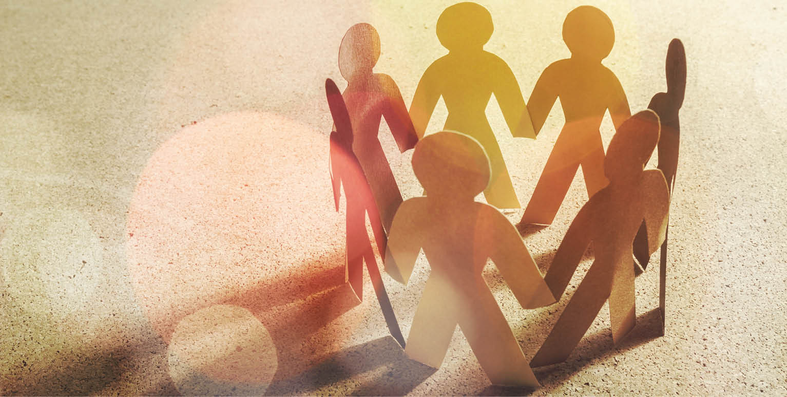 Paper cutout figures holding hands in a circle cast long shadows on a textured surface with warm sunlight creating lens flare effects. Description generated by AI